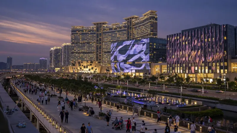 Crowds of people walk along a waterfront promenade at dusk in front of illuminated modern skyscrapers and buildings.